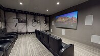 This private screening room is designed to deliver an immersive cinematic experience, combining thoughtful thematic design with high-quality audio-visual technology. The focal point of the space is a large wall-mounted display screen positioned on a neutral gray accent wall, flanked by flush-mounted in-wall speakers that form part of a comprehensive surround sound system. Additional speakers are integrated throughout the room, ensuring balanced audio coverage from every seat.

Seating is arranged in two tiered rows of black leather reclining chairs, each equipped with built-in cup holders, providing both comfort and a classic movie theater feel. The layout allows for clear sightlines to the screen from all positions.

The most distinctive design element is the full-scale film countdown mural that spans the rear and side walls, rendered in a vintage black-and-white aesthetic with film strip borders framing oversized countdown numerals. Wall-mounted sconce lighting accents the mural and contributes to the room's atmospheric ambiance. A bold red entry door with a porthole-style window serves as a striking contrast to the otherwise dark, moody interior.

The floor is finished with a textured, gray-toned carpet that complements the overall color palette. Recessed ceiling lighting provides practical illumination, completing this well-appointed, cinema-inspired space.