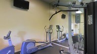 This single-level fitness room is designed to offer a practical and well-equipped workout space. The room features a selection of cardio equipment, including elliptical trainers positioned on dark rubber flooring that helps define the exercise area and provides cushioning underfoot. A cable weight machine anchored to the ceiling extends across the room, offering resistance training options for a variety of upper-body exercises. Wall-mounted flat-screen televisions are positioned at strategic heights, allowing users to stay entertained during workouts. A large mirror spans one wall, creating a sense of expanded space while allowing users to monitor their form during exercise. The walls are finished in a neutral, light tone that contributes to a clean and functional atmosphere. The overall layout is efficiently organized, maximizing the available floor space to accommodate multiple users simultaneously. The combination of cardio machines, strength training equipment, and entertainment options makes this fitness room a versatile facility suited for a range of workout routines.