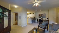 This single-floor residence offers a thoughtfully designed open-concept layout that seamlessly connects the main living areas. The living room is bright and inviting, featuring plush carpeting, a ceiling fan with integrated lighting, and recessed lights that contribute to a warm, comfortable ambiance. The space flows naturally into a dedicated dining area, which comfortably accommodates a full dining set — ideal for everyday meals or entertaining guests.

The kitchen is efficiently laid out with white cabinetry, white appliances, and a raised breakfast bar with seating for multiple bar stools, making it a central hub for both cooking and casual dining. Decorative greenery displayed atop the upper cabinets adds a finishing touch to the kitchen's clean aesthetic.

A partial wall elegantly separates the living zone from the kitchen, providing definition between the spaces while maintaining an open and airy feel throughout. Neutral tones in both the walls and flooring create a cohesive, understated palette that makes the space feel welcoming and versatile. A glimpse through a doorway reveals a private bathroom, completing this well-rounded and functional single-floor home.