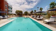 Located at 4201 Jamboree Road in Newport Beach, California, this outdoor amenity space is a thoughtfully designed resort-style deck serving a modern multi-story residential community. The grounds are organized into distinct zones that together create a cohesive, resort-inspired environment beneath the Southern California sky.

At the heart of the space is a large, sparkling swimming pool surrounded by a generous sun deck furnished with chaise lounges and shaded by classic navy market umbrellas. Mature palm trees and colorful plantings frame the perimeter, providing both privacy and a lush, tropical atmosphere.

A wide pedestrian walkway bisects the courtyard, connecting separate wings of the community and lined with sleek glass railings that preserve unobstructed views of the surrounding landscape and distant hills. Along this promenade, a linear fire feature filled with vivid blue glass stones serves as a striking focal point, flanked by built-in concrete seating and outdoor grilling stations ideal for entertaining.

A second distinct patio area features large-format tile pavers, wood deck accents, tall sculptural planters, and comfortable wicker lounge furniture arranged beneath pergola structures. The modern architecture of the surrounding buildings—characterized by clean lines, bold terracotta and grey paneling, and glass-enclosed balconies—provides an elegant backdrop throughout. The overall ambiance is polished, open, and inviting.
