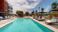 Located at 4201 Jamboree Road in Newport Beach, California, this outdoor amenity space is a thoughtfully designed resort-style deck serving a modern multi-story residential community. The grounds are organized into distinct zones that together create a cohesive, resort-inspired environment beneath the Southern California sky.

At the heart of the space is a large, sparkling swimming pool surrounded by a generous sun deck furnished with chaise lounges and shaded by classic navy market umbrellas. Mature palm trees and colorful plantings frame the perimeter, providing both privacy and a lush, tropical atmosphere.

A wide pedestrian walkway bisects the courtyard, connecting separate wings of the community and lined with sleek glass railings that preserve unobstructed views of the surrounding landscape and distant hills. Along this promenade, a linear fire feature filled with vivid blue glass stones serves as a striking focal point, flanked by built-in concrete seating and outdoor grilling stations ideal for entertaining.

A second distinct patio area features large-format tile pavers, wood deck accents, tall sculptural planters, and comfortable wicker lounge furniture arranged beneath pergola structures. The modern architecture of the surrounding buildings—characterized by clean lines, bold terracotta and grey paneling, and glass-enclosed balconies—provides an elegant backdrop throughout. The overall ambiance is polished, open, and inviting.