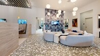 Located at 3068 Briarcliff Rd NE in Atlanta, GA, this elegantly designed single-floor community lounge offers a sophisticated and welcoming atmosphere. The space is anchored by a dramatic botanical-print feature wall that soars toward the high ceilings, creating a bold focal point in the main lounge area. Rounded, modular sofas in soft neutral tones are arranged atop a distinctive hexagonal tile floor, lending the room a curated, contemporary feel. Globe pendant lights hang at varying heights throughout, casting a warm, ambient glow that complements the abundant natural light streaming in through large floor-to-ceiling windows. A central bar island with a sleek countertop and warm amber bar stools serves as a functional gathering hub, framed by gold-toned exposed ceiling beams and marble-clad columns that add a refined, upscale touch. An adjacent dining area features velvet blue chairs and white marble-top tables set against an eye-catching arched wood slat wall with integrated backlighting, offering an intimate setting for smaller gatherings. A convenience retail nook with refrigerated displays adds everyday practicality. The overall ambiance is polished yet inviting, seamlessly blending luxury finishes with comfortable, functional design throughout.