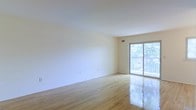 This single-floor space offers a clean, well-maintained interior defined by its light-filled and open character. The main living area is finished with polished light hardwood floors that reflect natural light beautifully, contributing to a warm and inviting atmosphere throughout. Smooth white walls provide a neutral backdrop, making the space feel airy and versatile.

A standout feature of the living area is the full-width sliding glass door that leads directly to a private balcony. The balcony is enclosed by a white railing and overlooks a lush, tree-lined backdrop, providing a serene outdoor retreat and bringing abundant natural light deep into the interior. The seamless connection between the indoor living space and the outdoor balcony enhances the overall sense of openness.

The ceiling is smooth and well-proportioned, complementing the overall brightness of the space. HVAC ventilation is integrated discreetly into the ceiling, ensuring climate comfort without detracting from the clean aesthetic. The overall design is simple and functional, with a neutral palette that allows for a wide range of interior styling possibilities. This space is well-suited for those who value natural light, outdoor connectivity, and a fresh, uncluttered living environment.