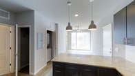 Located at 1420 Renaissance Drive in Longmont, Colorado, this single-floor residence offers a thoughtfully designed open-concept layout with modern finishes throughout. The kitchen serves as a central highlight, featuring a large island topped with light granite countertops and dark cabinetry with gold-toned hardware. Stylish pendant lights hang above the island, providing both functional task lighting and a contemporary aesthetic touch. The kitchen flows seamlessly into the living area, which is bathed in natural light from a large sliding glass door and window that invite the outdoors in, offering views of the surrounding area. A ceiling fan overhead adds to the comfort of the space. The unit also includes a dedicated in-unit laundry area with a stacked washer and dryer, offering added convenience. Light wood-look flooring runs throughout, complementing the clean, neutral color palette of the walls and ceilings. Recessed lighting and an electrical panel are also visible, reflecting a well-appointed and functional design. The overall ambiance is bright, clean, and contemporary, making this a comfortable and practical living space in the heart of Longmont, CO.