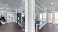 This single-story fitness facility is thoughtfully designed to support an active lifestyle. The space is finished with soft blue-gray walls, white crown molding, and dark flooring throughout, creating a clean and functional aesthetic.

The layout is divided into distinct zones. A built-in cubby and bench unit near the entrance provides convenient storage for personal belongings, with individual compartments and a lower bench for ease of use. Clipboard holders mounted on the wall suggest a space designed for organized, structured activity.

Adjacent to the entry area, a mirrored section features floor-to-ceiling mirrors along the walls, ideal for stretching, yoga, or group exercise. A stainless steel waste receptacle and a wall-mounted dispenser — likely for sanitizing wipes or paper towels — add practical touches to the space.

Beyond, the exercise area is visible, equipped with cardio machines including treadmills and a stair climber. Natural light filters in through multiple windows and a glass-paneled door, keeping the environment bright and inviting. Recessed lighting supplements the natural light throughout. The space is clean, efficiently organized, and well-suited for fitness use.