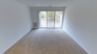 This single-floor residential space offers a clean, functional layout designed for comfortable everyday living. The main living area is a standout feature, characterized by its open, airy feel and neutral-toned carpeting that extends throughout the room. Large sliding glass doors serve as the focal point of the space, flooding the interior with natural light and providing direct access to a private balcony. The balcony overlooks a lush, wooded backdrop, offering a peaceful and natural outdoor view. A wall-mounted heating and ventilation unit is conveniently positioned beside the sliding doors, ensuring year-round climate comfort. The walls are finished in a crisp white, contributing to the bright and neutral ambiance that makes the space feel open and versatile. The textured ceiling adds a subtle architectural detail consistent with classic residential construction. Overall, the space presents a straightforward, well-maintained interior with practical amenities, ample natural light, and a seamless indoor-outdoor connection through its balcony access — making it a functional and inviting place to call home.