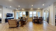 This single-floor space offers a thoughtfully designed open-concept layout that seamlessly blends the living and dining areas into one cohesive, inviting environment. The warm-toned hardwood flooring runs continuously throughout, anchoring the space with a sense of warmth and continuity.

The living area is comfortably appointed with upholstered sofas and a classic round wooden accent table, creating an ideal setting for relaxation or conversation. A wall-mounted television and framed artwork add functional and decorative touches to the space. Recessed lighting in the textured ceiling provides even, ambient illumination that complements the natural light streaming in through multiple large windows dressed with horizontal blinds.

The dining area is equally well-appointed, featuring a sturdy dark wood dining table surrounded by a generous set of mission-style chairs, making it well-suited for gatherings. A tall decorative plant adds a natural, refreshing element between the two areas, softening the transition while enhancing the overall ambiance.

The neutral wall tones and clean architectural lines lend the space a timeless, understated elegance. The abundance of windows along the exterior wall allows natural light to fill the room throughout the day, contributing to the bright and airy atmosphere that defines this well-balanced, functional space.