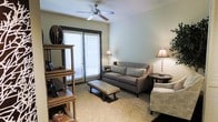 Located at 935 Garden Park Dr. in Allen, Texas, this single-floor residence offers a thoughtfully designed living space with a welcoming and comfortable ambiance. The living area serves as the heart of the home, featuring soft, neutral-toned walls that create a calm and inviting atmosphere. Plush carpeting runs throughout, adding warmth underfoot, while a ceiling fan overhead provides both style and practical comfort. A sliding glass door floods the room with natural light, framed by white horizontal blinds that allow for adjustable privacy and illumination. The space is well-proportioned, offering enough room for relaxed seating arrangements, including a sofa, accent chair, and a rectangular ottoman that doubles as a coffee table. Accent lamps on either side of the seating area contribute to a cozy, layered lighting scheme. A freestanding open shelving unit near the entrance adds both decorative and functional storage. The overall design aesthetic is clean and understated, with coordinated furnishings in soft grays and browns that complement the neutral palette of the space. The layout encourages comfortable everyday living while maintaining an organized and uncluttered feel throughout.