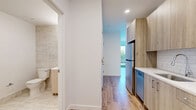 Located at 25 Maple Ave in New Rochelle, NY, this efficiently designed single-floor unit offers a thoughtfully laid-out interior with modern finishes throughout. Light wood-tone flooring runs through the main living and kitchen areas, creating a warm, cohesive aesthetic against the clean white walls.

The kitchen is a standout feature, outfitted with light wood cabinetry, white quartz countertops, a classic subway tile backsplash, and a full suite of stainless steel appliances including a range, over-the-range microwave, refrigerator, and dishwasher. A functional hallway connects the rooms, providing smooth circulation through the unit.

The main room is bright and airy, benefiting from abundant natural light streaming through large floor-to-ceiling windows and a glass door that opens to an outdoor area with lush greenery views. The space feels open and versatile, well-suited for both living and sleeping arrangements.

The bathroom features large-format marble-style tile on both the floors and walls, lending a clean, spa-like quality. It includes a toilet, vanity, and a generously sized tub and shower area with an accessible grab bar. Additional conveniences include two closets for ample storage and a dedicated laundry area, making this a highly functional and well-appointed unit.
