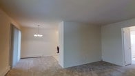 This single-floor space presents an open, functional layout characterized by neutral beige walls and wall-to-wall carpeting throughout. The interior is divided into connected zones by a partial wall partition, creating a natural separation between living areas while maintaining an open feel. In the adjoining room, a modest chandelier with multiple light fixtures serves as a central decorative element, adding a subtle touch of elegance to the otherwise understated space. The ceilings are smooth and standard in height, contributing to a clean, uncluttered aesthetic. Baseboard heating units are visible along the walls, indicating the primary source of climate control throughout the unit. A fire extinguisher is mounted near the partial dividing wall, reflecting attention to safety. The overall color palette is light and neutral, with beige tones dominating both the walls and flooring, creating a cohesive and calm ambiance. The space is currently unfurnished, allowing for flexible furniture arrangement and customization. The layout and design are straightforward and practical, suited for everyday living on a single level.