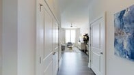 This single-floor residence at Overture offers a thoughtfully designed layout that balances functionality and comfort. A wide entryway corridor sets the tone upon entry, flanked by panel-style closet doors with sleek brushed nickel hardware, providing generous built-in storage. Dark hardwood-style flooring runs throughout, creating a warm contrast against the crisp white walls and trim.

The entryway opens into a well-proportioned living room bathed in natural light from multiple large windows dressed with elegant floor-length curtains. A ceiling fan with a brushed metal finish anchors the room from above, adding both style and practicality. The living space comfortably accommodates a sofa, accent chair, and coffee table arrangement centered on a decorative area rug, lending a relaxed yet polished atmosphere.

Along one wall, a sideboard credenza serves as an entertainment and décor focal point, complemented by accent lighting and mirrors. Lush greenery from an indoor plant adds a natural touch. The overall aesthetic is clean and contemporary, with a neutral palette of whites, grays, and soft blues that creates a cohesive, inviting environment throughout the space.