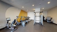 Located on Wilshire Boulevard in the heart of Los Angeles, this modern fitness center offers a well-equipped, single-floor workout facility designed to accommodate a comprehensive range of exercise routines. The space is divided into distinct functional zones, each thoughtfully arranged to maximize usability and flow.

The main cardio area features a row of treadmills and elliptical machines positioned along a boldly accented amber-yellow wall, creating an energizing atmosphere. Recessed lighting throughout the ceiling provides consistent, even illumination across the floor, which is finished with durable rubber flooring suited for high-traffic athletic use. A multi-functional cable and strength training machine stands centrally, offering users a versatile option for resistance-based workouts.

A separate strength training room houses several selectorized weight machines, including chest press, leg press, and other targeted resistance equipment, all set against clean, neutral-toned walls. This quieter zone is ideal for focused strength training sessions.

Near the entrance, large floor-to-ceiling windows flood the space with abundant natural light and provide views of the surrounding streetscape, including a glimpse of palm trees that evoke the quintessential Los Angeles setting. This area also includes a stationary bike and additional functional training equipment such as a squat rack and plyo boxes. The overall design is clean, contemporary, and purposefully laid out to support a full-body fitness regimen.