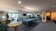 This single-floor clubroom and net café in Houston, TX presents a thoughtfully designed, multi-zone social space with a distinct contemporary aesthetic. The layout is anchored by a central game area housing a full-size billiard table beneath a coffered ceiling with gold-leaf accents and modern pendant lighting. A dramatic sculptural partition—composed of layered circular cutouts with a metallic, textured finish—divides the game zone from a spacious lounge area without fully enclosing either space, maintaining an open, airy flow throughout.

The lounge features generous sectional seating in muted blue-gray tones, accented with earth-toned throw pillows and centered around a dark wood coffee table. Adjacent seating clusters with yellow and neutral upholstered sofas add warmth alongside decorative consoles and lush tropical plants. A wall-mounted flat-screen television with geometric framing serves as a focal point in this area.

A semi-private meeting nook includes a round dining table, modern chairs, large plantation-shuttered windows that invite natural light, and ornate branch-style chandeliers beneath bold circular gold ceiling medallions. A bar area with bar-height seating and pendant lighting rounds out the space, creating a well-equipped social environment suitable for both relaxed gatherings and casual meetings.