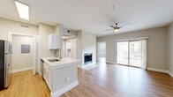 This single-floor residence offers a thoughtfully designed open-concept layout with warm hardwood-style flooring flowing throughout the entire space. The living area is a standout, centered around a white-tiled fireplace and fitted with a ceiling fan, creating a comfortable and inviting atmosphere. Sliding glass doors lead to a private balcony, allowing natural light to pour in and providing outdoor views. The kitchen is well-equipped with white cabinetry, a peninsula with a undermount sink and light quartz countertops, and stainless steel appliances including a refrigerator, range, and microwave. A dedicated hallway connects the kitchen to additional rooms, maintaining a practical flow throughout the unit. The bedroom is generously sized, featuring a ceiling fan, a large window with natural light, and double bi-fold closet doors offering ample storage. The bathroom includes a full bathtub and shower combo, white vanity cabinetry with brushed nickel hardware, and hardwood-style flooring. Neutral warm-gray wall tones are consistent throughout, contributing to a clean, cohesive aesthetic. The dollhouse view confirms a compact yet efficiently organized floor plan that maximizes every area of the space.