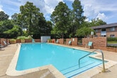 Located at 100 Lockwood Ct in Columbus, Georgia, this outdoor community pool area offers a well-appointed recreational space designed for relaxation and leisure. The centerpiece is a generously sized swimming pool with clear, inviting blue water, framed by a wide concrete pool deck that provides ample room for sunbathing and socializing. Turquoise lounge chairs are positioned throughout the deck, offering comfortable spots to unwind under the open sky.

A distinctive octagonal pavilion with a charming conical dark roof serves as a focal point of the space, accompanied by outdoor dining tables shaded by red umbrellas — ideal for gathering and outdoor meals. The pool area is enclosed by classic brick walls with ornamental iron fencing, lending the space a sense of privacy and security while maintaining an open, airy feel.

Lush mature trees and well-maintained greenery surround the perimeter, providing natural shade and a serene backdrop. Raised planters with ornamental grasses and flowering shrubs add thoughtful landscaping detail. The brick residential buildings visible in the background complement the traditional architectural character of the community. Bathed in natural sunlight beneath open skies, this outdoor pool amenity presents a welcoming and functional communal retreat.