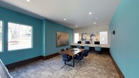 This well-appointed lounge offers a versatile environment suited for both group collaboration and individual productivity. The space is anchored by a rich wood-topped conference table surrounded by modern dark chairs, providing an ideal setting for meetings or group work sessions. Along one wall, a built-in countertop workstation accommodates multiple computer monitors, offering dedicated spaces for focused tasks. The design aesthetic strikes a balance between professionalism and comfort, highlighted by a bold teal accent wall that adds a pop of color and visual depth to the room. Complementing this are three coordinating abstract art pieces mounted above the workstation area, alongside a larger statement painting on the teal wall, contributing to the lounge's curated, contemporary ambiance. Recessed lighting throughout the ceiling ensures the space is well-lit, while natural light filters in through windows along the perimeter. The flooring features a subtly patterned carpet that ties the room's color palette together. Wall sconces flanking the artwork add warm accent lighting, enhancing the inviting atmosphere of this thoughtfully designed communal space.
