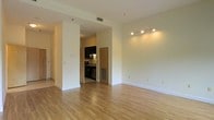 This single-floor residence offers a thoughtfully designed layout that balances open living space with functional utility. The main living area is expansive and airy, featuring warm hardwood flooring that extends throughout, crisp white walls, and track lighting that provides focused illumination across the room. The neutral color palette creates a bright, welcoming ambiance with a clean, minimalist aesthetic.

A defining architectural feature is the partial wall partition that separates the living area from the kitchen, providing a sense of division while maintaining an open, connected feel. The kitchen is outfitted with dark cabinetry that creates a striking contrast against the lighter tones of the surrounding space. It includes modern appliances, a sink with task lighting above, and a practical layout designed for everyday use. A utility vent is also incorporated into the kitchen area, suggesting efficient climate control throughout the unit.

Ceiling-mounted HVAC vents are visible in the living area, ensuring year-round comfort. The smooth, high ceilings contribute to the overall sense of spaciousness. The corridor visible along one side hints at additional rooms beyond the main living space. Overall, this residence presents a tidy, well-maintained interior that is move-in ready with a straightforward, functional design.