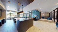 This elegantly designed single-floor amenity space combines contemporary aesthetics with functional comfort throughout its open, flowing layout. The main lobby and lounge area is anchored by large-format tile flooring and floor-to-ceiling black-framed windows that flood the interior with natural light and provide sweeping views of an outdoor pool courtyard. A prominent reception or bar counter features a dark stone surface, white subway tile base, and dramatic brass globe pendant lighting suspended from a wood-paneled ceiling. The accompanying kitchenette area showcases rich dark cabinetry, teal mosaic tile backsplash, and a generous white countertop with upholstered bar seating in coordinating teal tones. The lounge is furnished with a curated mix of leather sofas, teal velvet accent chairs, and built-in gray banquette seating paired with marble-topped side tables and sculptural brass chandeliers. Warm wood column accents and bold abstract artwork contribute to the space's refined atmosphere. A dedicated conference or business room features herringbone hardwood flooring, a glass-top table with nailhead-trim chairs, built-in shelving, a wall-mounted display, and a steel-framed interior window. Throughout the space, thoughtful lighting, rich materials, and a cohesive teal-and-brass color palette create a polished, welcoming environment.