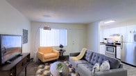 This single-floor space features an open-concept layout that connects the living area to the kitchen, creating a cohesive and functional flow throughout. The living area is well-lit, benefiting from a large window dressed with vertical blinds that allow generous natural light to fill the room. The textured ceiling adds a subtle architectural detail overhead, while dark hardwood-style flooring grounds the space with a warm contrast to the neutral wall tones.

The living area is generously proportioned, comfortably accommodating a tufted gray sofa, an accent armchair in a warm mustard tone, a round coffee table, and a patterned area rug — all contributing to a modern, mid-century inspired aesthetic. A stylish arc floor lamp provides additional ambient lighting.

Through an open passageway, the kitchen is visible, featuring white cabinetry and dark countertops, suggesting a clean and practical cooking space. The overall ambiance of this residence is bright, airy, and inviting, with a neutral palette that provides a versatile backdrop adaptable to a variety of interior design preferences.