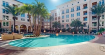 This upscale residential complex boasts a Mediterranean-inspired aesthetic with its pastel-colored facades and arched windows. The multi-story buildings encircle a meticulously landscaped courtyard, creating a serene oasis for residents. At the heart of the complex lies a expansive, crystal-clear swimming pool, its curved edges mirroring the architectural elements of the surrounding structures. The pool area is flanked by numerous lounge chairs, perfect for sunbathing or relaxation.

The outdoor spaces are adorned with a variety of palm trees, providing both shade and a tropical ambiance. Lush gardens with diverse plant life, including succulents and ornamental grasses, add texture and color to the environment. Brick pathways weave through the grounds, connecting various seating areas furnished with comfortable outdoor furniture, including plush sofas and dining sets.

For added comfort, the complex features covered cabanas and pergolas, offering shaded retreats from the sun. Fire pits and water features contribute to the luxurious atmosphere, creating focal points for social gatherings. The overall design seamlessly blends indoor and outdoor living, with many apartments featuring balconies overlooking the picturesque courtyard. This resort-like setting provides residents with a tranquil escape and a multitude of amenities for recreation and relaxation.