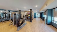 This fitness center is a comprehensively outfitted workout facility designed to support a wide range of exercise routines. The space is organized into distinct functional zones, each catering to different training needs. The cardio area features a row of treadmills and elliptical machines positioned along one wall, with large mirrors reflecting natural light from the windows and amplifying the sense of openness throughout the room. Recessed ceiling lighting provides consistent illumination across the entire floor.

The strength training section is anchored by a multi-station cable machine from Precor, complemented by a weight bench and additional resistance equipment. A dedicated dumbbell rack offers a full range of free weights, ideal for targeted muscle training. Structural columns define the space without interrupting the overall flow between zones.

A well-stocked storage unit displays an assortment of stability balls and medicine balls in various sizes, supporting functional fitness and flexibility training. The flooring transitions between light wood hardwood in the open areas and rubberized mat surfaces beneath the cardio equipment, providing both comfort and safety. Teal accent walls add a vibrant, energizing aesthetic to the otherwise neutral color palette, creating a motivating environment for users of all fitness levels.