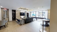 This single-floor space is designed as a contemporary communal lounge, combining sophisticated aesthetics with functional comfort. The room is anchored by a striking marble-style accent wall that serves as a backdrop for a large wall-mounted flat-screen television, flanked by dark built-in bookshelves that add depth and character to the space. The seating area features a plush gray sofa and modern accent chairs arranged atop a area rug, creating an inviting gathering zone. Two elegant multi-bulb chandelier fixtures hang from a recessed tray ceiling, providing warm, ambient lighting that enhances the upscale atmosphere. Light wood-toned flooring runs throughout, adding warmth and contrast to the room's cooler tones. Along the far wall, expansive floor-to-ceiling windows and glass panels flood the interior with natural light while offering views of an outdoor patio area. A high-top bar counter with stools is positioned near the windows, ideal for casual seating or socializing. The overall ambiance is polished and modern, balancing open, airy spaces with carefully curated design elements suited for relaxation or social gatherings.
