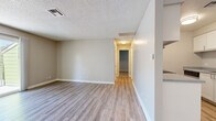 This single-floor unit offers a clean, functional layout with a cohesive neutral color palette throughout. The open living area is finished with light wood-style vinyl plank flooring and features sliding glass doors that lead directly to a private balcony, allowing natural light to fill the space and providing pleasant outdoor views. The living area connects seamlessly to the kitchen, which is equipped with white cabinetry, a stainless steel refrigerator, a range, and a granite-style countertop. A stacked washer and dryer unit is conveniently located within the kitchen area, adding in-unit laundry functionality. A hallway with ceiling-mounted lighting connects the main living spaces to the private rooms. The bedroom features plush wall-to-wall carpeting and a window fitted with blinds that brings in natural light. The full bathroom includes a tub-shower combination, a toilet, a vanity with a granite-style countertop, and wood-style flooring. Throughout the unit, textured ceilings, neutral beige walls, and clean white trim create a consistent and tidy aesthetic. The layout, as seen in the overhead dollhouse view, is compact yet efficiently organized, making practical use of every area within the space.