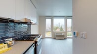 Located at 910 SW 18th Ave in Portland, Oregon, this single-floor unit features a contemporary design with a clean, neutral palette and light wood-style flooring throughout. The open layout connects the kitchen and living areas seamlessly, creating a functional and cohesive living space.

The kitchen is well-appointed with stainless steel appliances, including a gas range, dishwasher, and refrigerator. White flat-front cabinetry extends to the ceiling, maximizing storage, while a striking dark geometric hex tile backsplash adds a bold design accent. A dedicated range hood and built-in microwave round out the culinary setup.

The living area is bathed in natural light from large floor-to-ceiling windows that frame elevated views of the Portland cityscape and surrounding greenery, offering an impressive urban panorama. A wall-mounted HVAC unit provides climate control directly within the living space.

The bathroom features a full soaking tub with a clean, floor-to-ceiling white tile surround and chrome fixtures, offering a crisp, modern aesthetic. A hallway with recessed lighting connects the spaces efficiently, with built-in storage adding practical utility. The overall ambiance is bright, minimalist, and modern, well-suited to urban living in Portland's westside neighborhood.