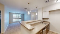 This single-floor unit presents a thoughtfully designed open-concept layout connecting the kitchen and living areas. The kitchen is equipped with stainless steel appliances, including a refrigerator, microwave, and range, complemented by a peninsula with a built-in sink and light countertops. White upper cabinetry and warm wood-toned lower cabinets provide ample storage, while light hardwood-style flooring grounds the space. Pendant lighting above the peninsula adds a contemporary touch.

The living area extends from the kitchen, featuring plush carpeting, track lighting, and a large window that admits generous natural light and outdoor views. The open floorplan model reveals multiple bedrooms, each carpeted and well-lit with windows. Bedroom sizes offer comfortable proportions with easy access to the hallway.

Both bathrooms are finished consistently with tile flooring in a stone-like pattern, dark wood vanities with white countertops, and walk-in showers. One bathroom includes grab bars, indicating accessible design considerations. Large mirrors above the vanities enhance the sense of space in each bathroom. The overall aesthetic is clean, neutral, and functional throughout.