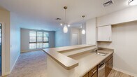 This single-floor unit presents a thoughtfully designed open-concept layout connecting the kitchen and living areas. The kitchen is equipped with stainless steel appliances, including a refrigerator, microwave, and range, complemented by a peninsula with a built-in sink and light countertops. White upper cabinetry and warm wood-toned lower cabinets provide ample storage, while light hardwood-style flooring grounds the space. Pendant lighting above the peninsula adds a contemporary touch.

The living area extends from the kitchen, featuring plush carpeting, track lighting, and a large window that admits generous natural light and outdoor views. The open floorplan model reveals multiple bedrooms, each carpeted and well-lit with windows. Bedroom sizes offer comfortable proportions with easy access to the hallway.

Both bathrooms are finished consistently with tile flooring in a stone-like pattern, dark wood vanities with white countertops, and walk-in showers. One bathroom includes grab bars, indicating accessible design considerations. Large mirrors above the vanities enhance the sense of space in each bathroom. The overall aesthetic is clean, neutral, and functional throughout.