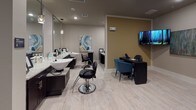 This well-appointed hair salon offers a complete range of beauty services within a thoughtfully designed, single-floor layout. The space features light wood-style flooring throughout, creating a clean and cohesive aesthetic that ties each area together seamlessly.

The primary styling area is anchored by sleek, dark-cabinetry stations topped with granite-style countertops and large mirrors, each illuminated by elegant pendant lighting. Professional hydraulic styling chairs are positioned at each station, providing a functional and polished setting for hair services. Dedicated shampoo basins with accompanying dryer chairs are set against a bold teal accent wall, which is adorned with a series of fashion illustration artworks, adding a chic, artistic focal point to the space.

A separate nail and pedicure area features a cushioned massage pedicure chair alongside a manicure desk with teal upholstered seating, set against warm tan-toned walls. A wall-mounted flat-screen television enhances the client experience in this section. Recessed lighting throughout ensures consistent, even illumination across all service areas.

Overall, the salon presents a modern yet welcoming ambiance, balancing professional functionality with stylish interior design elements that create a comfortable environment for both clients and practitioners.