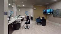 This well-appointed hair salon offers a complete range of beauty services within a thoughtfully designed, single-floor layout. The space features light wood-style flooring throughout, creating a clean and cohesive aesthetic that ties each area together seamlessly.

The primary styling area is anchored by sleek, dark-cabinetry stations topped with granite-style countertops and large mirrors, each illuminated by elegant pendant lighting. Professional hydraulic styling chairs are positioned at each station, providing a functional and polished setting for hair services. Dedicated shampoo basins with accompanying dryer chairs are set against a bold teal accent wall, which is adorned with a series of fashion illustration artworks, adding a chic, artistic focal point to the space.

A separate nail and pedicure area features a cushioned massage pedicure chair alongside a manicure desk with teal upholstered seating, set against warm tan-toned walls. A wall-mounted flat-screen television enhances the client experience in this section. Recessed lighting throughout ensures consistent, even illumination across all service areas.

Overall, the salon presents a modern yet welcoming ambiance, balancing professional functionality with stylish interior design elements that create a comfortable environment for both clients and practitioners.