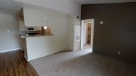 This single-floor residential unit offers a practical and comfortable layout designed for everyday living. The main living area is finished with light-colored carpeting, creating a warm and inviting atmosphere. A pass-through counter connects the living space to the kitchen, allowing for an open feel while maintaining a defined cooking area. The kitchen is equipped with built-in appliances, including a microwave and refrigerator, and features overhead cabinetry for ample storage.

The living space is accented by a contrasting dark feature wall, adding a modern touch to the otherwise neutral color palette of whites and light tones throughout. Vaulted or elevated ceilings contribute to a sense of openness in the space. Ventilation is well-considered, with visible HVAC registers integrated into the walls and ceiling.

A doorway from the main living area leads to a bedroom, which includes access to a walk-in closet, offering generous storage space. The overall design is clean and functional, with a cohesive neutral aesthetic that provides a versatile backdrop for a variety of interior styles. This unit is well-suited for those seeking a straightforward, move-in-ready living space with a practical floor plan.