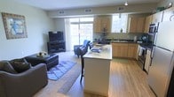 This single-floor unit showcases an open-concept layout that seamlessly connects the kitchen, dining area, and living room. The kitchen is outfitted with stainless steel appliances, including a gas range, built-in microwave, and refrigerator, complemented by light maple cabinetry and dark countertops. Track lighting illuminates the space overhead, while a central island with a light-toned countertop doubles as a breakfast bar with seating for two, making it ideal for casual dining or entertaining.

Adjacent to the kitchen, the dining area accommodates a two-person high-top table, flowing naturally into the living room. The living space benefits from large windows that invite abundant natural light and provide access to a balcony, enhancing the overall sense of openness. Light hardwood-style flooring extends throughout the main areas, transitioning to carpet in the living room.

A dedicated laundry area is conveniently tucked near the kitchen, housing a full-sized washer and dryer. The warm, neutral color palette and cohesive design elements throughout contribute to a comfortable and functional living environment.