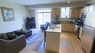 This single-floor unit showcases an open-concept layout that seamlessly connects the kitchen, dining area, and living room. The kitchen is outfitted with stainless steel appliances, including a gas range, built-in microwave, and refrigerator, complemented by light maple cabinetry and dark countertops. Track lighting illuminates the space overhead, while a central island with a light-toned countertop doubles as a breakfast bar with seating for two, making it ideal for casual dining or entertaining.

Adjacent to the kitchen, the dining area accommodates a two-person high-top table, flowing naturally into the living room. The living space benefits from large windows that invite abundant natural light and provide access to a balcony, enhancing the overall sense of openness. Light hardwood-style flooring extends throughout the main areas, transitioning to carpet in the living room.

A dedicated laundry area is conveniently tucked near the kitchen, housing a full-sized washer and dryer. The warm, neutral color palette and cohesive design elements throughout contribute to a comfortable and functional living environment.