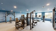 Located at 601 Sayre Ave in Perth Amboy, NJ, this fitness center offers a generously sized, single-floor layout designed to support a full range of workout routines. The gym is finished with neutral carpet flooring and painted in a calming blue palette, with coordinating blue baseboards that lend the space a clean, cohesive aesthetic. Recessed lighting throughout ensures consistent, even illumination across the entire floor.

The main gym area is comprehensively outfitted with a wide variety of equipment. Strength training options include a multi-station cable machine, a power rack with bench, a dumbbell rack with a full set of free weights, and several selectorized resistance machines targeting different muscle groups. Cardio equipment lines the windowed wall, where multiple treadmills, an elliptical, a stationary bike, and a rowing machine are arranged to take advantage of the natural light and outdoor views. The windows along this wall provide ample daylight and a connection to the surrounding neighborhood.

The open floor plan allows for generous circulation space between equipment stations. Two bathrooms serve the facility, providing convenient amenities for users. The overall layout is functional and well-organized, making this fitness center a practical and fully equipped space for a broad range of exercise needs.