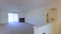 This single-floor space presents a straightforward and functional layout centered around a generously sized living area. The room is finished with wall-to-wall carpeting in a neutral tone, providing a comfortable and cohesive foundation throughout. Smooth, light-colored walls contribute to a clean and understated aesthetic, while a ceiling-mounted HVAC vent indicates a central air system for year-round climate control.

A built-in fireplace serves as a focal point along one wall, adding warmth and a welcoming element to the living space. Adjacent to the fireplace, a sliding glass door fitted with vertical blinds allows natural light to fill the room and provides access to an outdoor area. A partial white partition wall visible in the foreground suggests a defined transitional zone, possibly separating the living area from a dining or entryway space. A hallway visible in the background hints at additional rooms beyond. The overall ambiance is calm and neutral, offering a blank canvas well-suited for a variety of interior styles and furnishing arrangements.