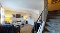 This two-story residence offers a well-organized layout distributed across two levels, connected by a carpeted staircase with a black metal railing. The main level centers around a living area finished with dark hardwood-style flooring, neutral-toned walls, and a textured ceiling. Natural light filters in through a window dressed with bold striped curtains, creating a warm and inviting atmosphere. The living space flows naturally into a separate dining area, which features the same flooring and is accented by decorative wall art and sunburst mirrors.

The galley-style kitchen is equipped with warm wood cabinetry, dark granite-style countertops, black appliances including a refrigerator and electric range, and wood-look flooring. A window above the sink allows natural light to brighten the space. A hallway connects the kitchen to additional rooms on the main level.

The upper level houses the bedrooms, each finished with plush carpet and neutral walls. The bedrooms receive natural light through windows with sheer curtains and blinds. A full bathroom on this level features a tub-shower combination with a curtain, hardwood-style flooring, and clean, simple finishes throughout.