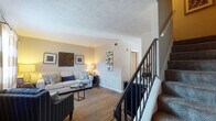 This two-story residence offers a well-organized layout distributed across two levels, connected by a carpeted staircase with a black metal railing. The main level centers around a living area finished with dark hardwood-style flooring, neutral-toned walls, and a textured ceiling. Natural light filters in through a window dressed with bold striped curtains, creating a warm and inviting atmosphere. The living space flows naturally into a separate dining area, which features the same flooring and is accented by decorative wall art and sunburst mirrors.

The galley-style kitchen is equipped with warm wood cabinetry, dark granite-style countertops, black appliances including a refrigerator and electric range, and wood-look flooring. A window above the sink allows natural light to brighten the space. A hallway connects the kitchen to additional rooms on the main level.

The upper level houses the bedrooms, each finished with plush carpet and neutral walls. The bedrooms receive natural light through windows with sheer curtains and blinds. A full bathroom on this level features a tub-shower combination with a curtain, hardwood-style flooring, and clean, simple finishes throughout.