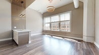 Located in the heart of downtown Dayton at 115 E Third St, this 2-bedroom, 1-bathroom unit blends modern design with urban character. The open-concept kitchen and living area serves as the centerpiece of the home, featuring wood-look flooring, soaring ceilings, and large windows that flood the space with natural light and offer views of the surrounding cityscape. A striking circular pendant chandelier anchors the living area, while the kitchen impresses with two-tone cabinetry, a decorative mosaic tile backsplash, stainless steel appliances, and a waterfall-edge marble countertop island with a gold-finish faucet. A hallway with wall sconce lighting connects the main living area to the private quarters. Both bedrooms are well-proportioned, each with hardwood-style flooring, neutral tones, and generous natural light from city-facing windows. The primary bedroom includes a spacious closet with dark wood bifold doors, and one bedroom features an architectural column detail that nods to the building's historic roots. The bathroom is finished with large-format marble-look tile on both the floors and tub surround, complete with a frameless sliding glass shower door for a clean, contemporary aesthetic. Three closets throughout the unit provide ample storage.