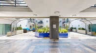Located at 3324 Castle Heights Ave in the Palms neighborhood of Los Angeles, this ground-floor common area serves as the central outdoor courtyard of a multi-story residential building. The space is defined by its expansive stone tile flooring, featuring a mix of earthy and cool-toned slate tiles that lend a sophisticated, durable finish throughout the walkways and gathering areas.

A partially covered corridor runs along the perimeter, providing shade while maintaining an open, airy feel. Architectural arched pergola structures add visual interest and frame the outdoor lounge area, which is furnished with contemporary rattan seating accented in vibrant blue upholstery — creating a welcoming social space for residents.

Lush landscaping is integrated throughout the courtyard via bold blue raised planters filled with drought-tolerant greenery, including succulents and ornamental grasses, complementing the Southern California climate. Clean white rectangular planters with snake plants line the quieter passageways, adding a minimalist, modern touch.

A centrally located utility column with secured access anchors the courtyard, while a pair of sleek, dark-doored elevators provides convenient vertical access to upper residential floors. The surrounding multi-story building features balconies that overlook the courtyard, creating a sense of enclosure and community. Abundant natural light floods the space, enhanced by the clear Los Angeles sky visible above.