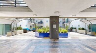 Located at 3324 Castle Heights Ave in the Palms neighborhood of Los Angeles, this ground-floor common area serves as the central outdoor courtyard of a multi-story residential building. The space is defined by its expansive stone tile flooring, featuring a mix of earthy and cool-toned slate tiles that lend a sophisticated, durable finish throughout the walkways and gathering areas.

A partially covered corridor runs along the perimeter, providing shade while maintaining an open, airy feel. Architectural arched pergola structures add visual interest and frame the outdoor lounge area, which is furnished with contemporary rattan seating accented in vibrant blue upholstery — creating a welcoming social space for residents.

Lush landscaping is integrated throughout the courtyard via bold blue raised planters filled with drought-tolerant greenery, including succulents and ornamental grasses, complementing the Southern California climate. Clean white rectangular planters with snake plants line the quieter passageways, adding a minimalist, modern touch.

A centrally located utility column with secured access anchors the courtyard, while a pair of sleek, dark-doored elevators provides convenient vertical access to upper residential floors. The surrounding multi-story building features balconies that overlook the courtyard, creating a sense of enclosure and community. Abundant natural light floods the space, enhanced by the clear Los Angeles sky visible above.