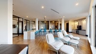 Located in Davenport, FL, this single-floor amenity space delivers a well-appointed, contemporary environment designed for both productivity and relaxation. The open-plan kitchen and living area anchors the space, featuring wood-look tile flooring, slate-blue cabinetry, a granite-topped wet bar, a beverage refrigerator, and large black-framed glass doors that flood the room with natural light and provide views of an outdoor patio with lush landscaping and palm trees. An expansive long white countertop bar with stylish seating connects the kitchen zone to the main lounge, where white swivel lounge chairs and teal accent tables create a bright, inviting gathering area. Black-framed glass partitions lend an open yet defined quality to the layout. The dining room is anchored by a substantial dark wood table paired with natural wood chairs, a wall-mounted display, and warm wood cabinetry — all beneath a deep teal accent ceiling. Three private offices provide focused work areas, each individually styled with bold patterned wallcovering, built-in wood desks, and blue task chairs. The connecting hallway features the same wood-look tile flooring and clean white walls, maintaining design continuity throughout. Large gridded windows throughout the space maximize natural light and offer outdoor views.