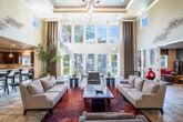 Located at 2311 Ivy Hill Way in San Ramon, CA, this community clubhouse is a well-appointed, single-floor facility designed to serve as a central hub for residents. The standout feature is the grand lounge area, distinguished by soaring double-height ceilings, decorative columns, and an elegant chandelier. A sweeping wall of tall windows and French doors floods the space with natural light while offering views of the surrounding palm-lined exterior. Neutral tones, stone tile flooring, and a vibrant area rug anchor the seating arrangement, creating an inviting and sophisticated atmosphere. Adjacent to the main lounge is a casual sitting room with a sectional sofa, accent windows, and direct access to an outdoor patio dining area — perfect for informal gatherings. The fully equipped kitchen features rich dark-wood cabinetry, granite countertops, stainless steel appliances, and ample storage, making it well-suited for catered events or communal cooking. A dedicated business center with workstations and recessed lighting provides a quiet, functional workspace. The entry foyer, accented with curated artwork and stone tile floors, sets an elegant tone upon arrival. Throughout, the design balances contemporary style with practical community amenities.