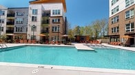 Located at 2312 Bristol Creek Dr in Morrisville, NC, this expansive outdoor amenity space is designed to deliver a resort-style experience within a vibrant residential community. At its centerpiece is a large, sparkling pool with an adjoining sun shelf, surrounded by a generous concrete deck lined with orange lounge chairs and shaded umbrella tables — ideal for relaxing under the North Carolina sun.

A covered outdoor lounge area provides a comfortable retreat, featuring exposed wooden beam ceilings, a sleek stainless steel linear fireplace set into a brick accent wall, and a wall-mounted flat-screen television — creating an inviting space for year-round gathering. Adjacent to this area, an outdoor kitchen and grilling station with bar-height seating offers a dedicated space for casual dining and entertaining.

The courtyard opens onto a well-maintained open lawn, framed by manicured landscaping and garden beds, with the multi-story residential buildings forming an architecturally cohesive backdrop in a mix of brick, blue-gray siding, and contemporary paneling. Ornamental black iron fencing delineates the pool and patio zones, adding a refined aesthetic touch. The overall ambiance is social, bright, and well-appointed, offering residents a comprehensive outdoor living experience.