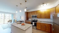 This single-floor residence offers a thoughtfully designed open-concept layout that seamlessly connects the kitchen, dining, and living areas. The kitchen is a standout feature, showcasing warm honey-toned cabinetry, granite countertops, a central island with an undermount sink, and a full suite of stainless steel appliances including a range, microwave, and refrigerator. Elegant pendant lighting hangs above the island, complementing the track lighting overhead and the light wood-style flooring that flows throughout the main living areas. The living room is bright and inviting, with neutral walls and a large sliding glass door that draws in natural light. Two bedrooms provide comfortable retreats, each featuring plush carpet, large windows with blinds, and ample access to closet space. The bathrooms are finished with granite-topped vanities and warm wood cabinetry consistent with the rest of the home. One bathroom includes a full shower-tub combination with clean tile surround, while the second serves as a well-appointed half bath. Throughout the space, a cohesive color palette of warm neutrals and crisp whites creates a calm, welcoming ambiance that is both functional and refined.