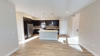 This single-floor residence offers a thoughtfully designed open-concept layout that maximizes both functionality and style. The kitchen serves as the heart of the space, showcasing rich dark espresso cabinetry that contrasts beautifully with light, neutral walls and warm hardwood-style flooring. A central island provides additional prep space and a natural gathering point, while pendant lighting above adds a modern decorative touch. Stainless steel appliances, including a refrigerator, range, and microwave, lend a clean, contemporary feel, complemented by a classic tile backsplash behind the range. Recessed ceiling lighting throughout ensures the space is well-illuminated, and ample natural light streams in, brightening the open floor plan. The seamless flow between the kitchen and the surrounding living areas creates a spacious, airy ambiance well-suited for both everyday living and entertaining. The neutral color palette and clean architectural lines give the space a timeless, move-in-ready appeal. Overall, the residence combines modern finishes with practical design, resulting in a comfortable and stylish environment.
