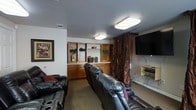 This single-floor space is thoughtfully designed as a dedicated home theater and entertainment room. The focal point is a large wall-mounted flat-screen television, positioned for optimal viewing from rows of plush black leather recliner sofas equipped with built-in cup holders — a nod to the classic cinema experience. The seating is arranged to ensure clear sightlines to the screen, making it an ideal setup for movie nights or watching sports.

Built-in wooden cabinetry lines the back wall, offering a combination of open shelving and closed lower cabinets. The shelving displays decorative greenery, small figurines, and other accents, adding warmth and personality to the space. A rich, dark curtain panel adds a dramatic, theater-like ambiance and helps manage light within the room.

The walls are painted in a clean, neutral tone, complemented by carpet flooring that enhances acoustics and comfort underfoot. Flush-mounted ceiling light fixtures provide even, practical illumination throughout. A framed movie-themed artwork on the wall further reinforces the cinematic theme of the space. Overall, the room strikes a balance between functional entertainment design and comfortable, inviting décor.