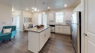 This single-floor residence offers a thoughtfully designed layout with multiple bedrooms, two full bathrooms, a contemporary kitchen, and dedicated utility space. The open-concept kitchen is a standout feature, showcasing crisp white shaker cabinetry, a central island with a gray quartz countertop and bar seating, stainless steel appliances including a refrigerator, range, microwave, and dishwasher, and pendant lighting overhead. Dark hardwood-style flooring runs throughout the kitchen and dining area, adding warmth and contrast to the bright, neutral palette. Natural light filters in through multiple windows fitted with white horizontal blinds. The bedrooms are carpeted and generously sized, each offering a calm, neutral atmosphere. One bedroom features a wide window with open outdoor views, while another is appointed with a dark-framed headboard and framed wall art. Both bathrooms are finished with white shaker vanities, gray countertops, dark wood-style flooring, and shower-tub combinations. A dedicated utility room houses an in-unit washer and dryer alongside a water heater, offering added convenience. Throughout the space, a consistent design language of white walls, neutral tones, and modern finishes creates a clean and cohesive living environment.
