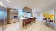 Located at 1517 Cameron Samuel Lane in West Palm Beach, Florida, this sleek, single-floor communal kitchen and lounge space offers a well-appointed environment designed for both functionality and social gathering. The open layout accommodates a fully equipped modern kitchen alongside a comfortable seating area, making it ideal for shared use.

The kitchen is outfitted with high-end stainless steel appliances, including a full-size refrigerator, wall ovens, and a built-in microwave, all set against a striking marble-patterned backsplash that extends to accent columns throughout the space. Taupe cabinetry with a sleek finish provides ample storage, while a generous white countertop runs along the perimeter, offering abundant workspace. A large picture window allows natural light to flood the kitchen area, providing a view of the greenery outside.

A prominent central island with a white countertop and warm wood base serves as the focal point of the space, flanked by stylish bar stools that invite casual dining and conversation. A substantial overhead range hood adds an industrial-chic element to the design.

The lounge area is anchored by high-top bistro tables paired with contemporary wooden bar chairs, arranged beneath vibrant large-scale citrus-themed artwork that injects bold color and energy into the otherwise neutral palette. Light wood-look tile flooring unifies the entire space, complementing the clean, modern aesthetic throughout.